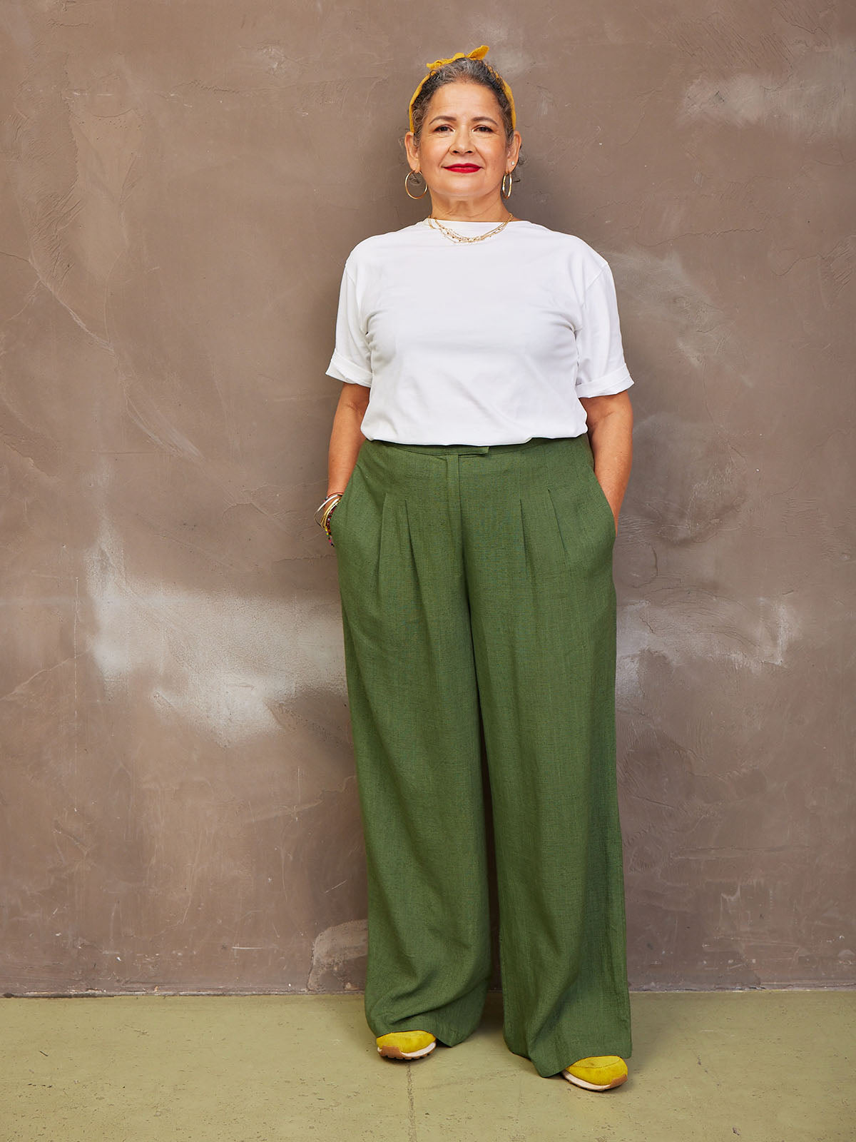 A person in a striped sweater and green pants stands against a textured wall.