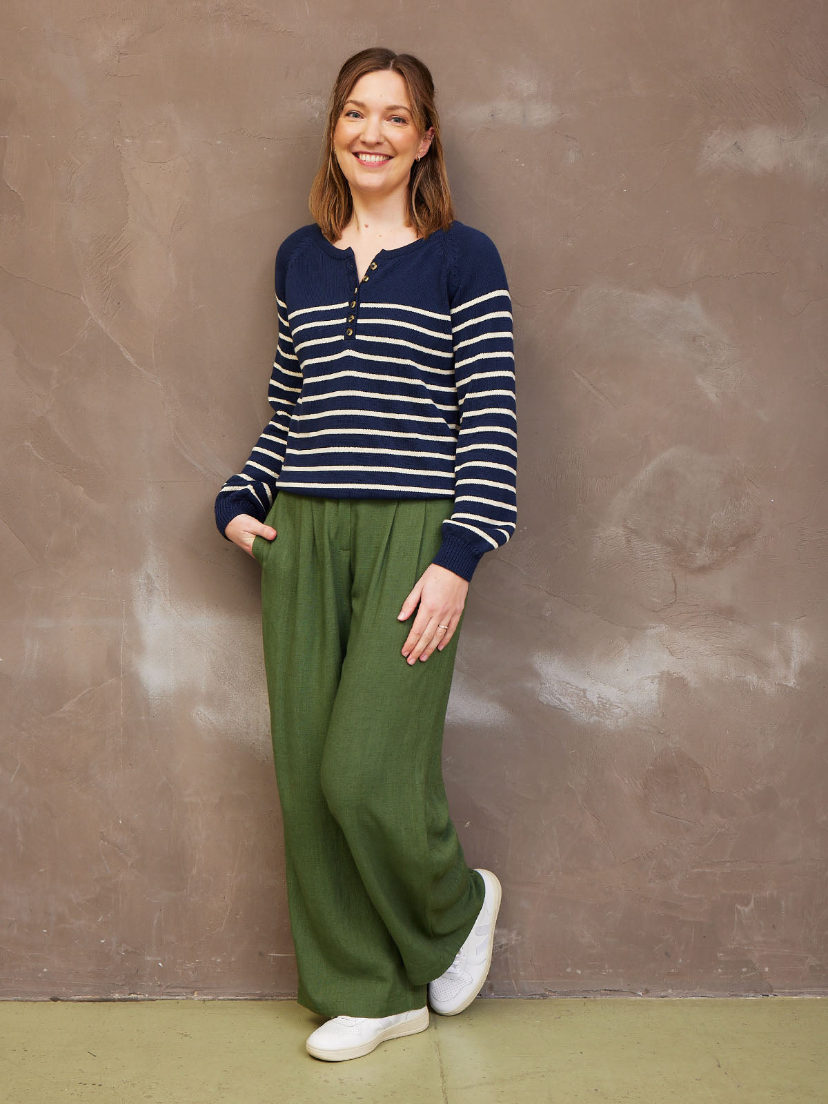 A person wearing a striped sweater and green pants, leaning against a textured wall.