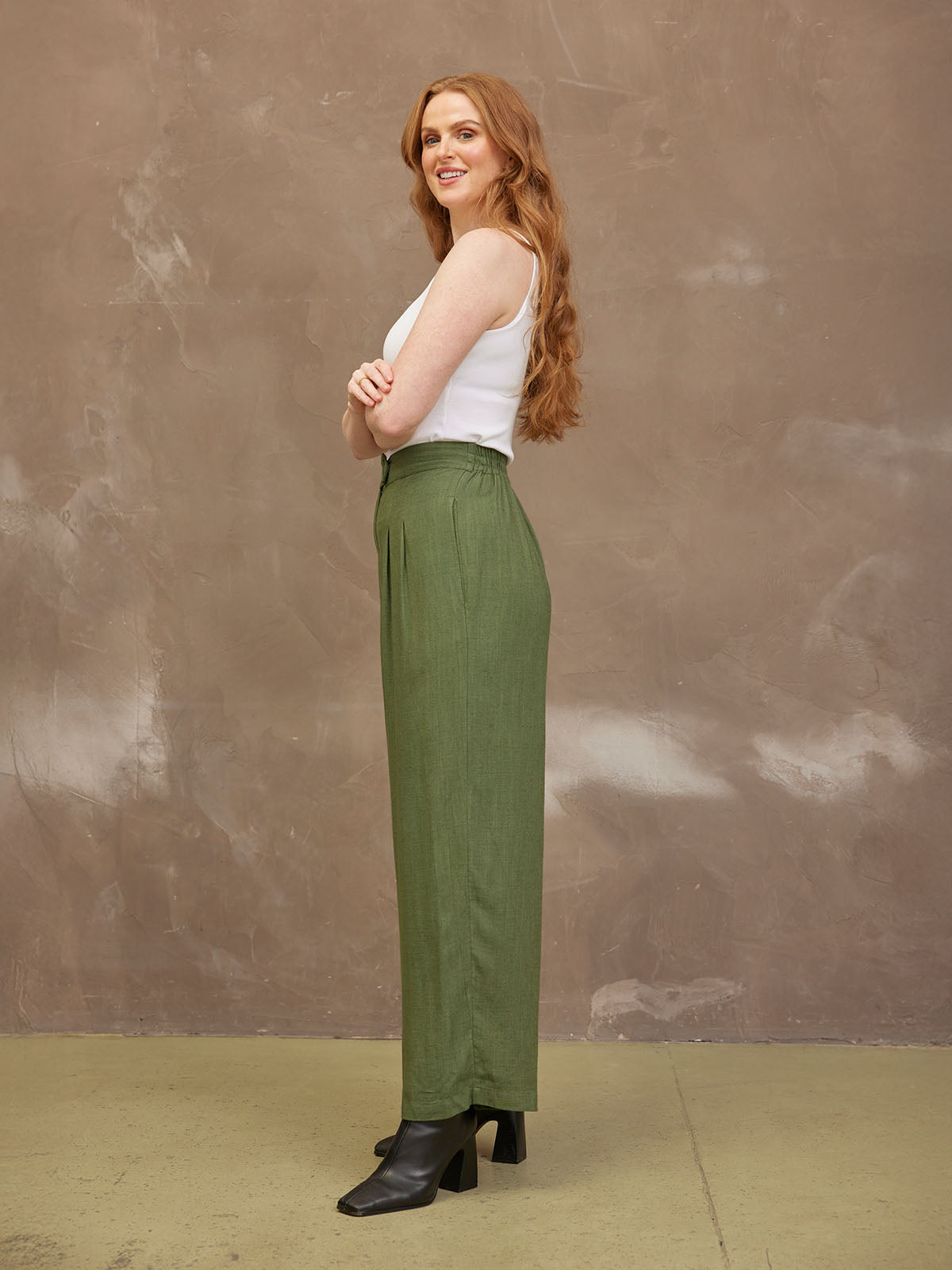 Woman in green pants and white top, smiling against a textured brown background.