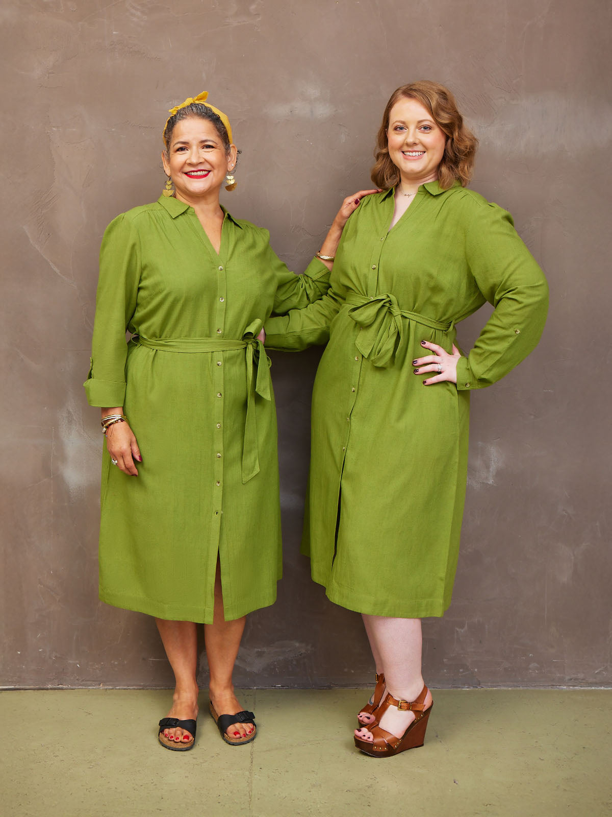 Two women smiling, wearing matching green dresses, standing against a neutral background.