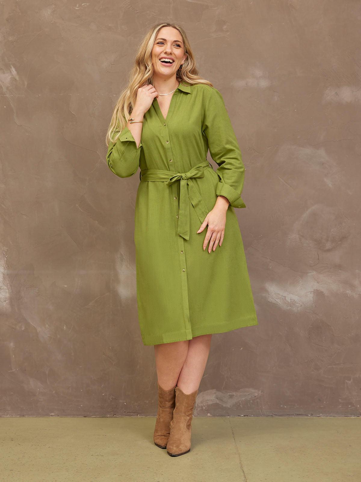 A woman in a green dress and brown boots, smiling against a textured brown background.