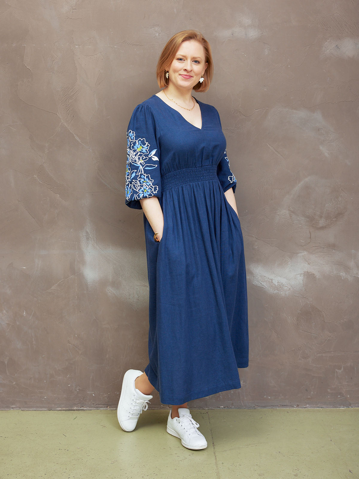 Person wearing a blue dress with floral embroidery and white sneakers, standing against a textured wall.