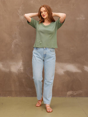 A smiling model with her eyes closed with both her hands behind her head, wearing the sustainable Lucy khaki t-shirt paired with jeans and sandals.