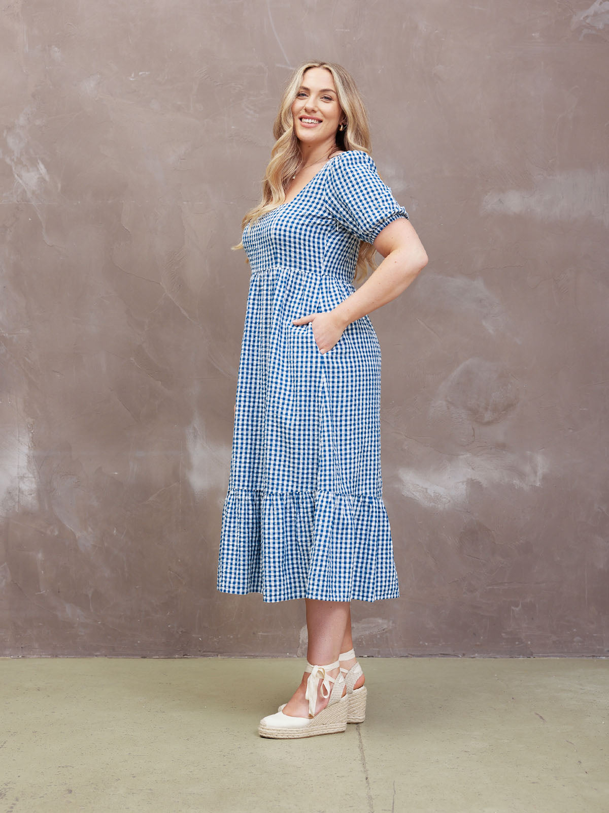 Smiling woman in a blue gingham dress standing against a textured brown wall.