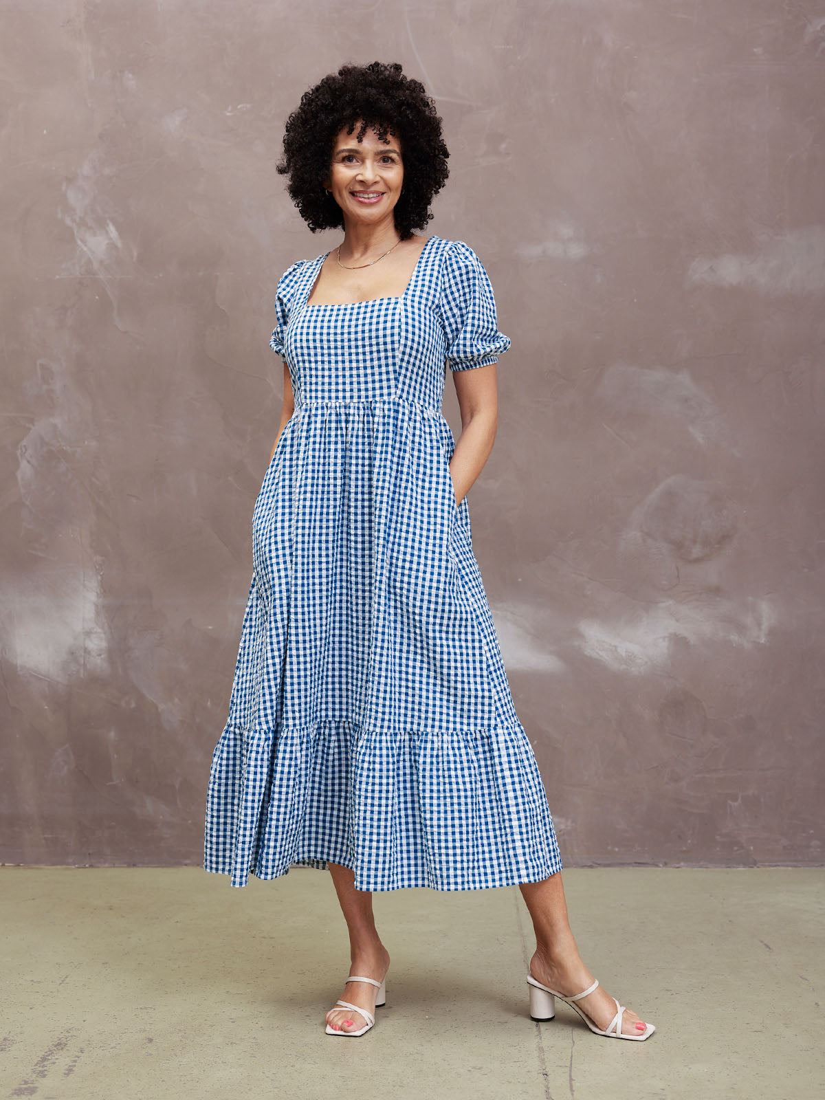 A person wearing a blue checkered dress and white sandals poses indoors.