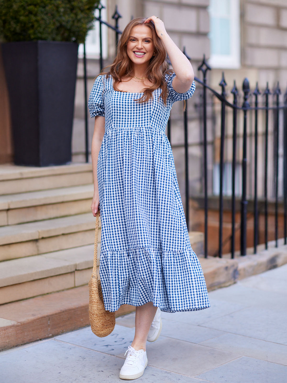 Woman in a blue gingham dress with a straw bag, smiling outdoors.