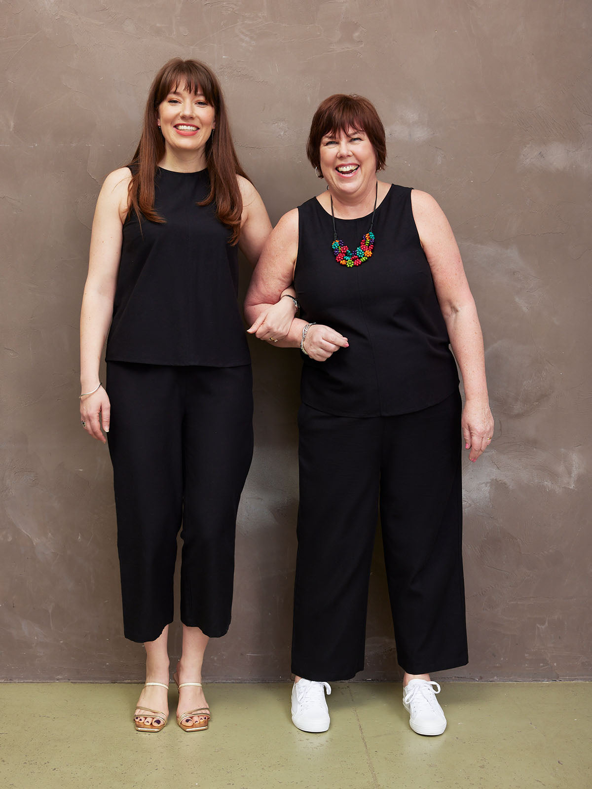 Two women in black outfits smiling against a brown wall.