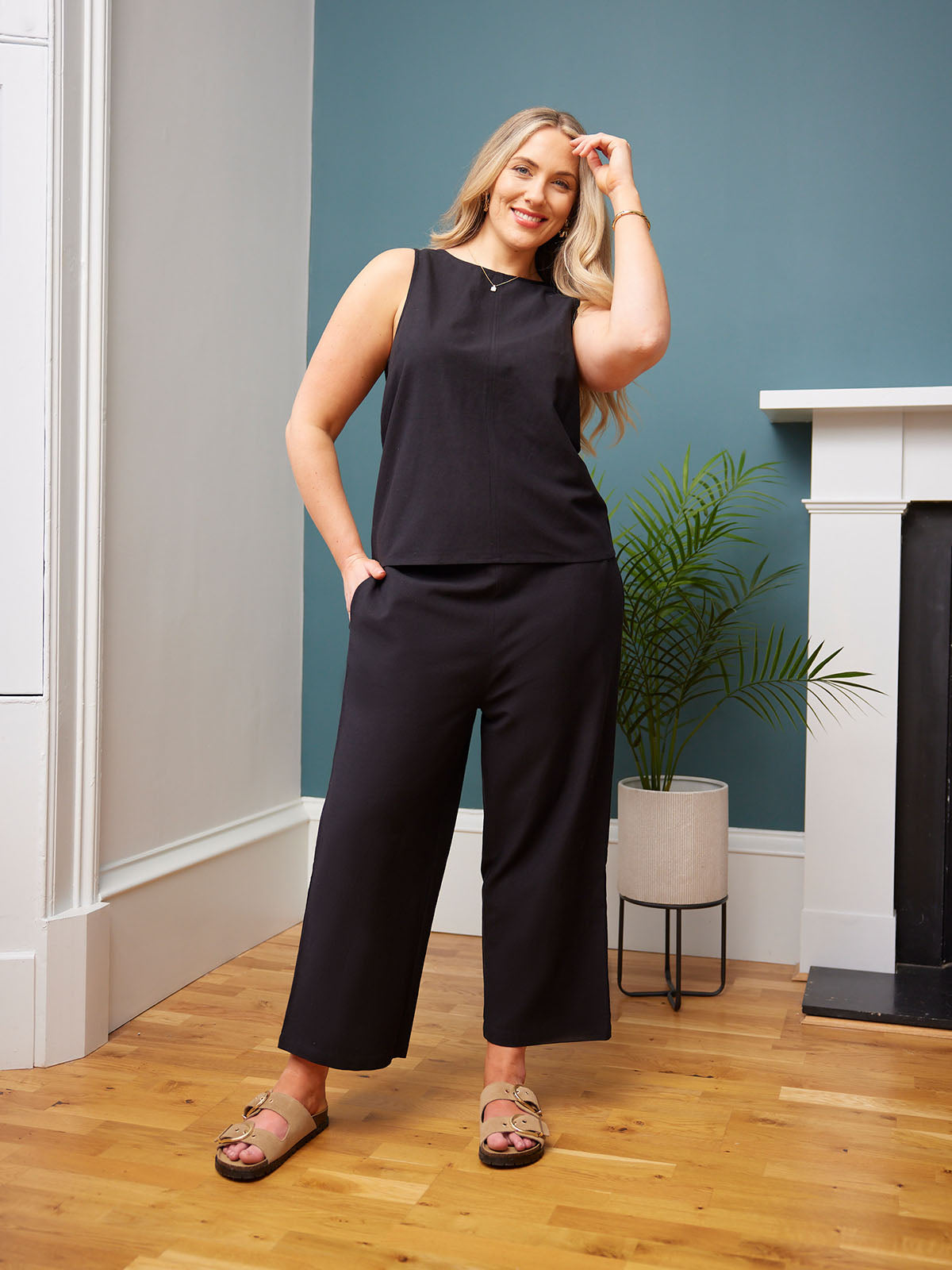 Person in a sleeveless black outfit and sandals, standing in a stylish room.