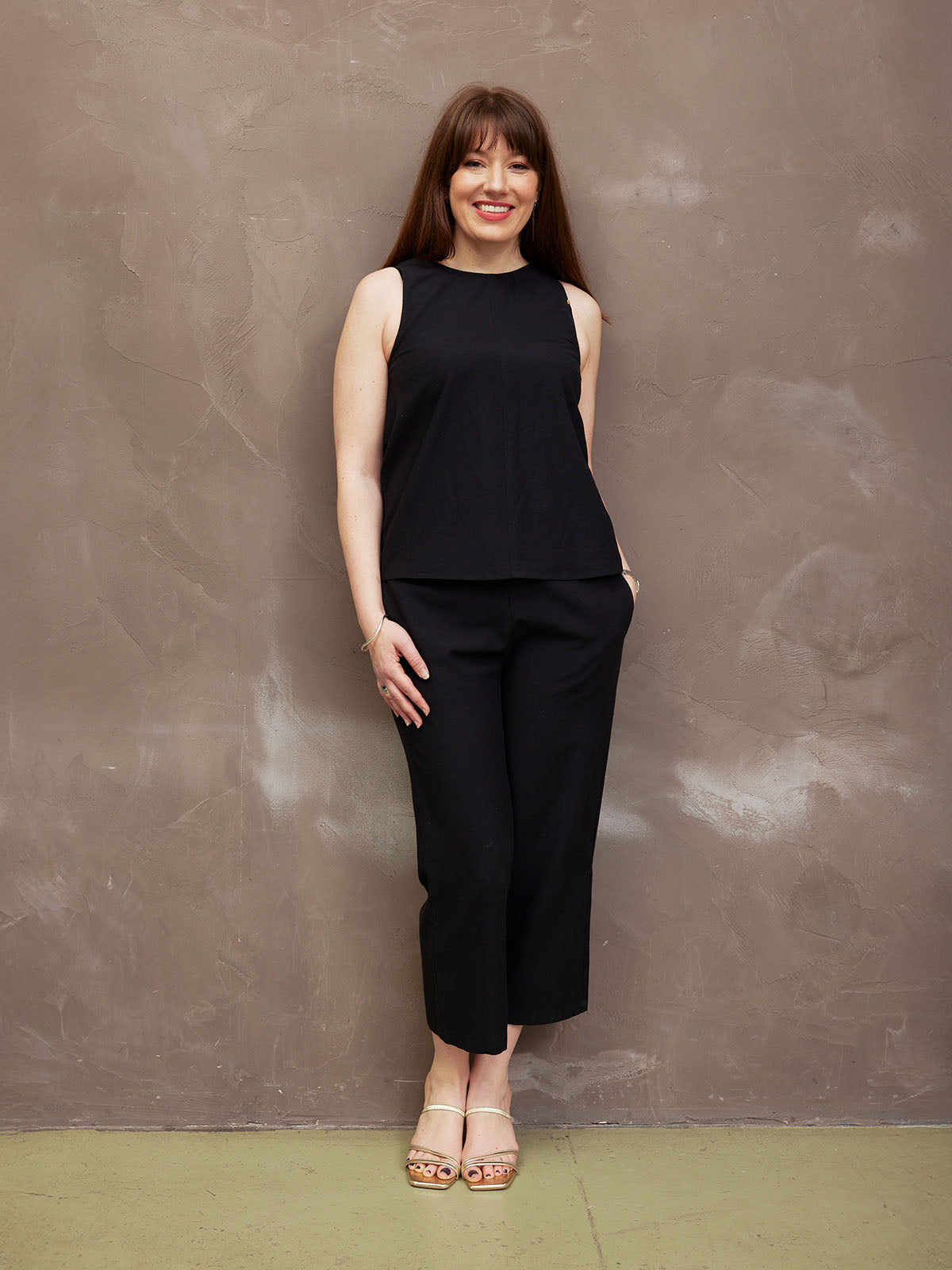 A woman in a black outfit stands smiling against a textured wall.