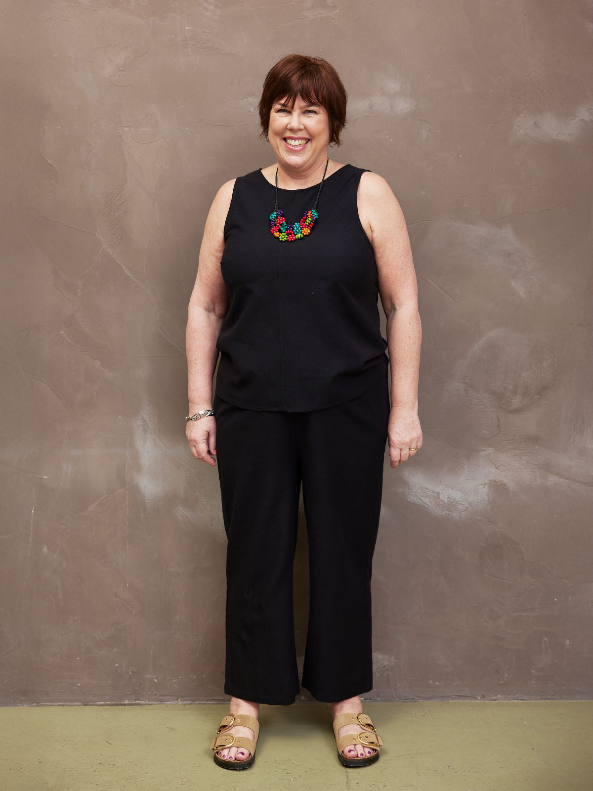 A person wearing a black outfit and colorful necklace, standing against a textured brown wall.