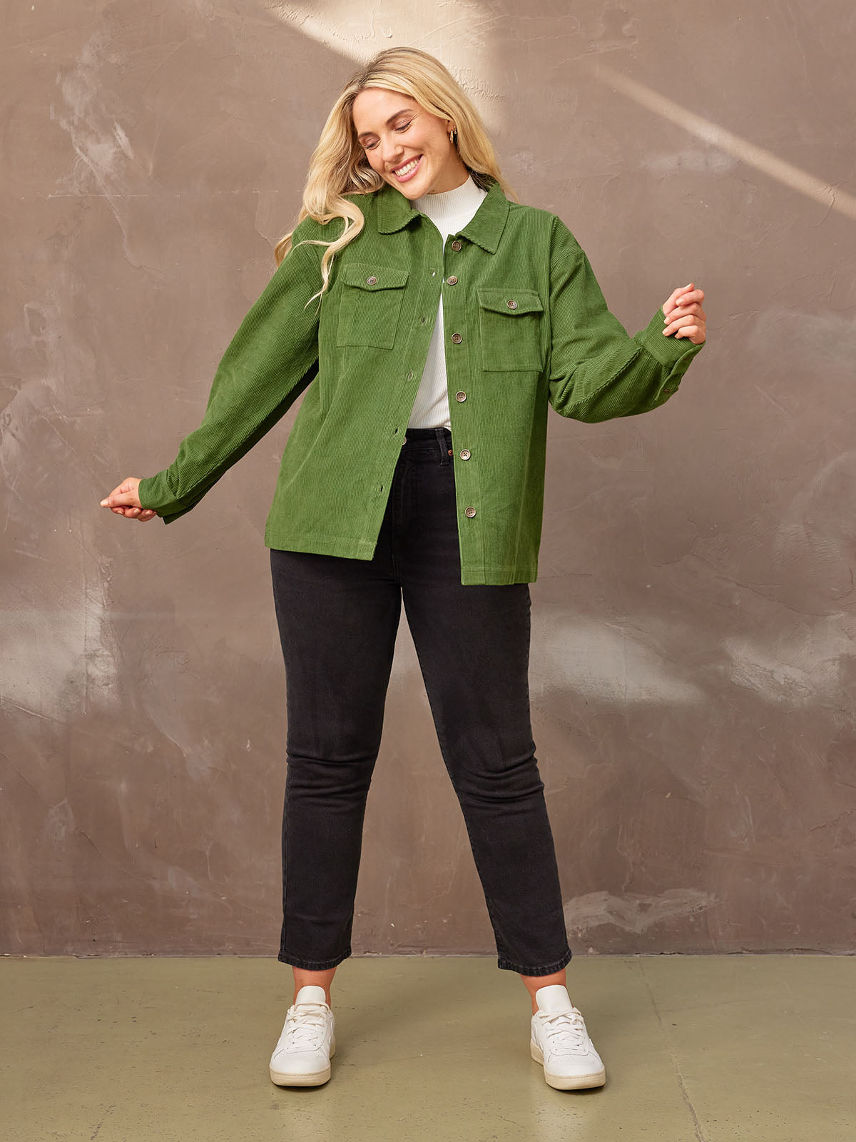 Smiling model in a cheerful pose with her arms out, in a sustainable olive coloured corduroy Yasmynn shacket paired with a white top and black trousers., looking at the ground.