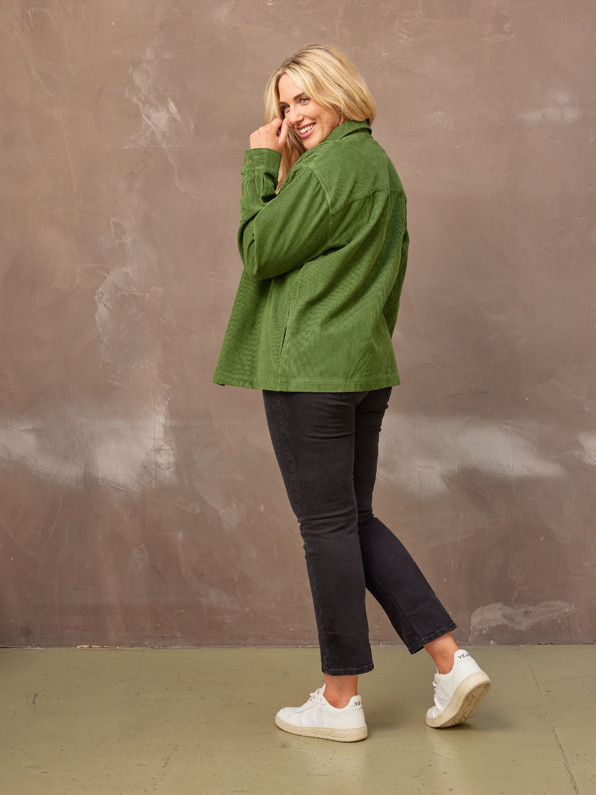 Sideview of a smiling model wearing the sustainable corduroy Yasmynn shacket in olive, paired with a white top and black trousers, with her left hand close to her face.