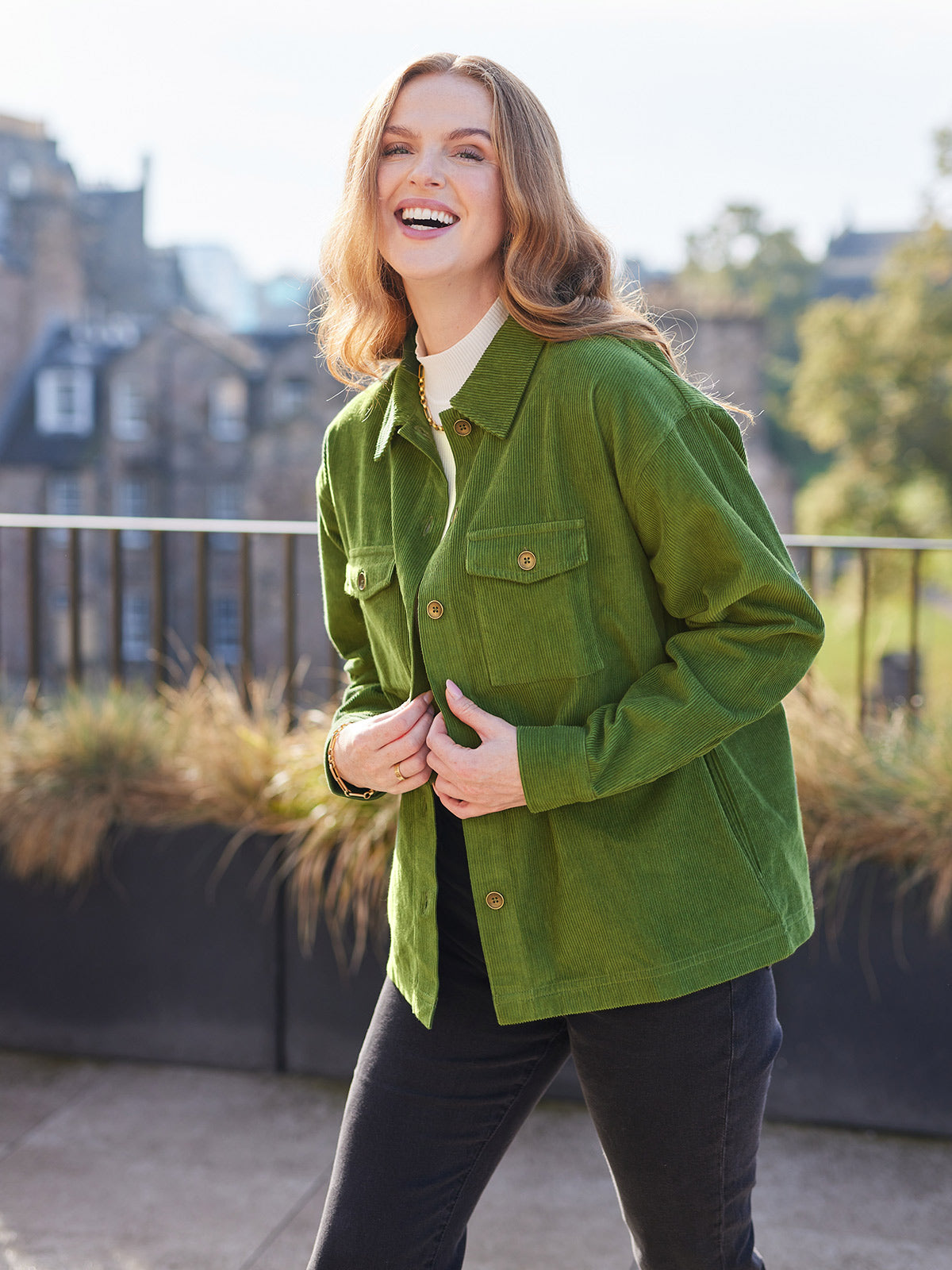 Smiling model looking at the camera and leaning forward, wearing the sustainable Yasmynn shacket in olive, paired with a white top and black trousers. Pictured in front of buildings outside with natural light.