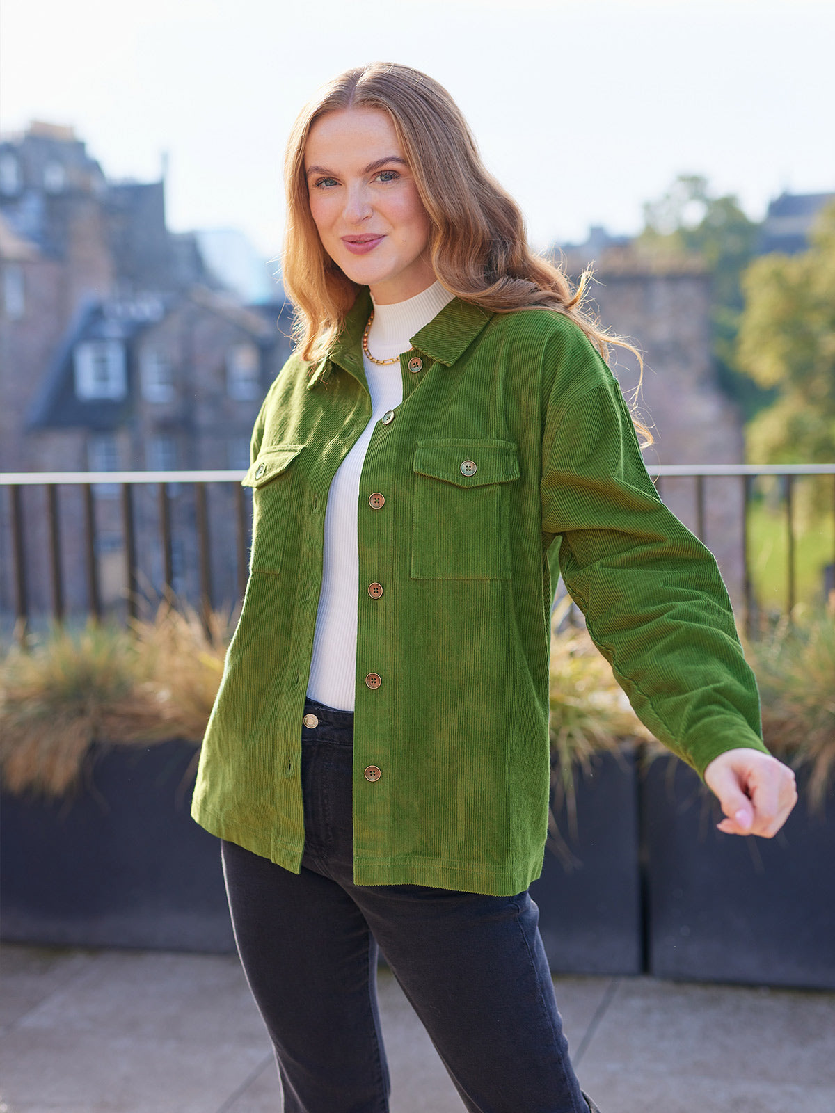 A model looking at the camera wearing the sustainable Yasmynn shacket in olive, paired with a white top and black trousers. Pictured in front of buildings outside with natural light.