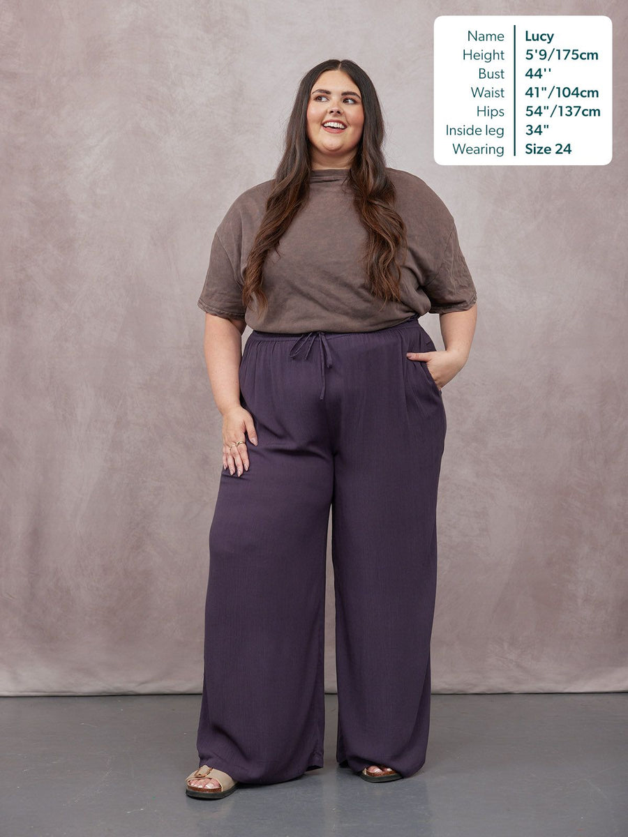 Model wearing wide-leg purple trousers with a drawstring waist, paired with a brown oversized tee and sandals.