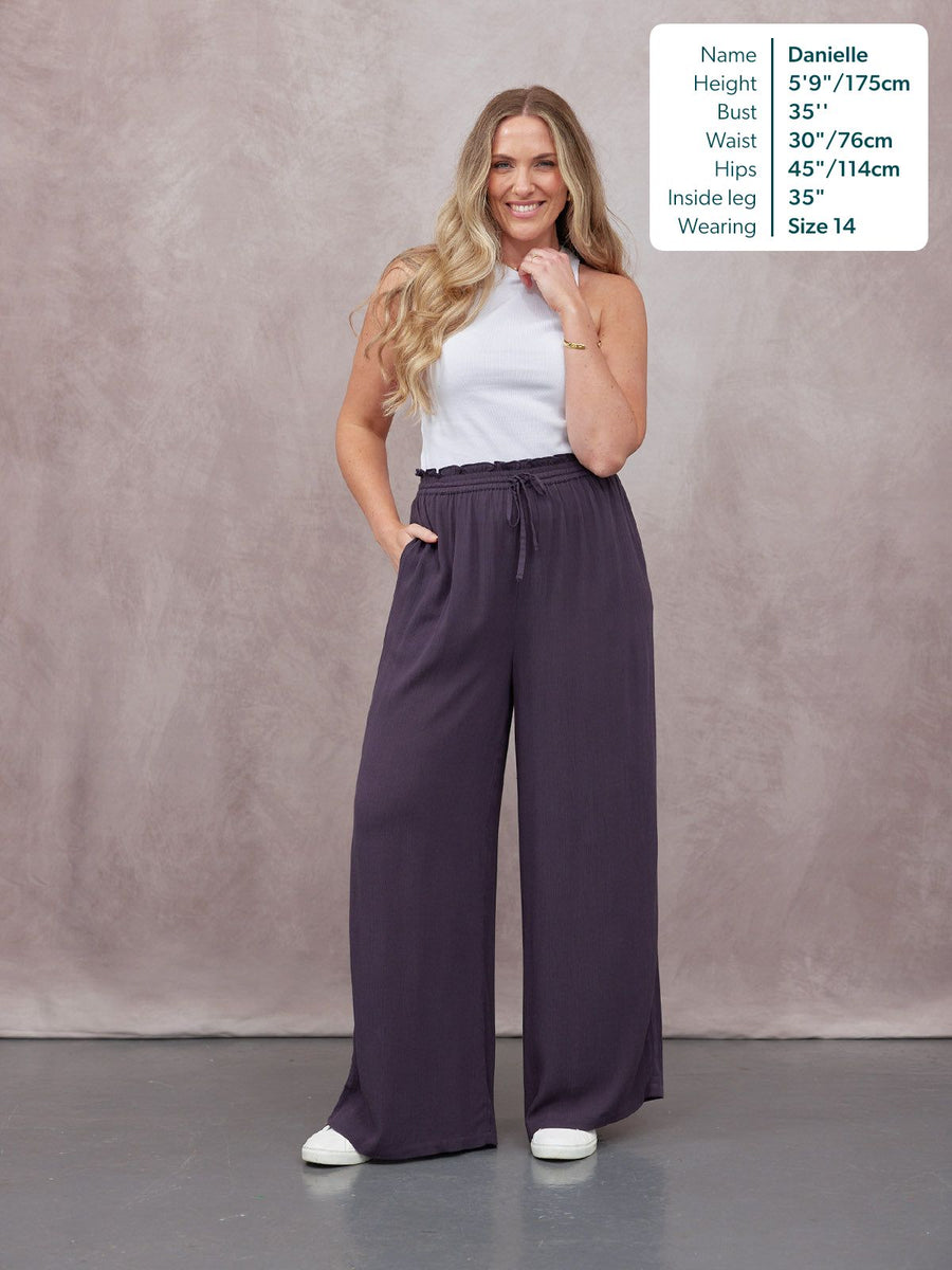 Model wearing dark purple wide-leg trousers with a drawstring waist and a white sleeveless top, paired with white trainers.