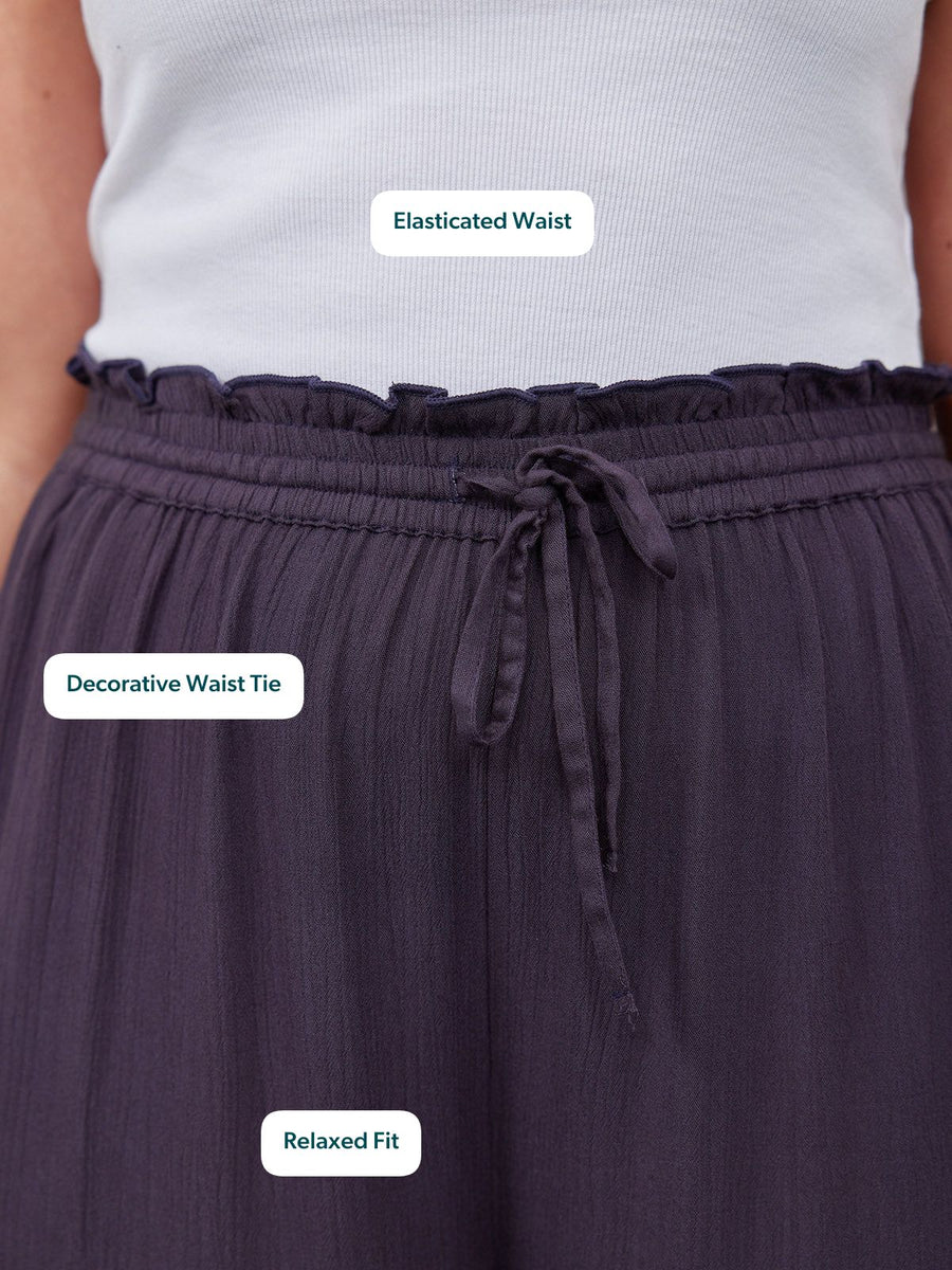 Close-up of navy wide-leg trousers with a ruffled elasticated waist and decorative tie, worn by a model with a white ribbed…