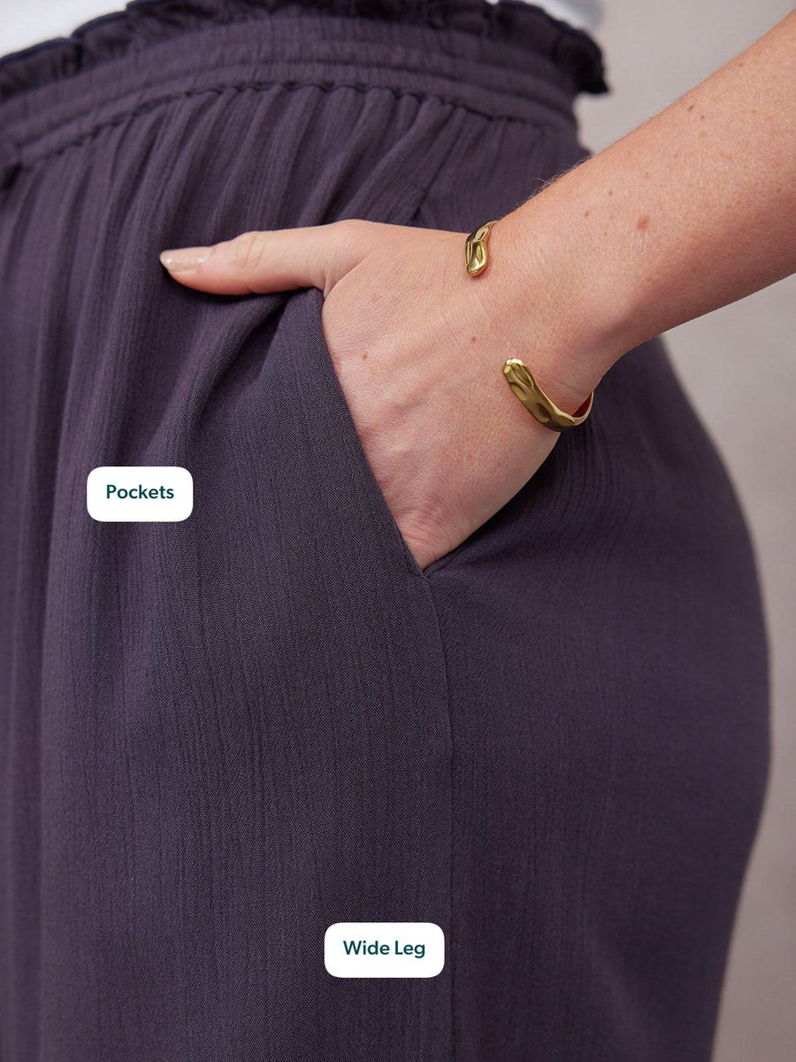 Close-up of dark purple wide-leg trousers with ruched waistband, side pocket, and gold cuff bracelets on model's wrist.