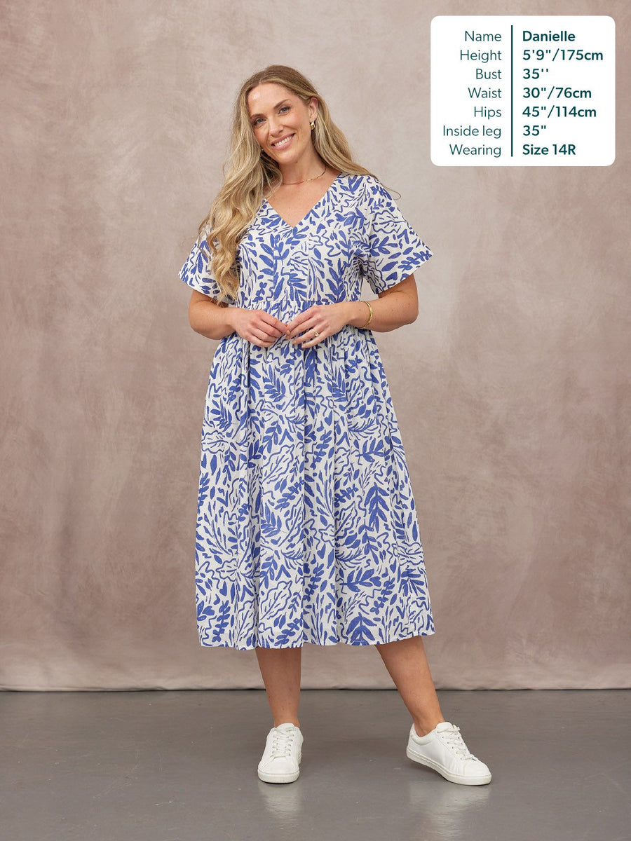 Model wearing a white and blue leaf print midi dress with flutter sleeves and a V-neckline, styled with white trainers.