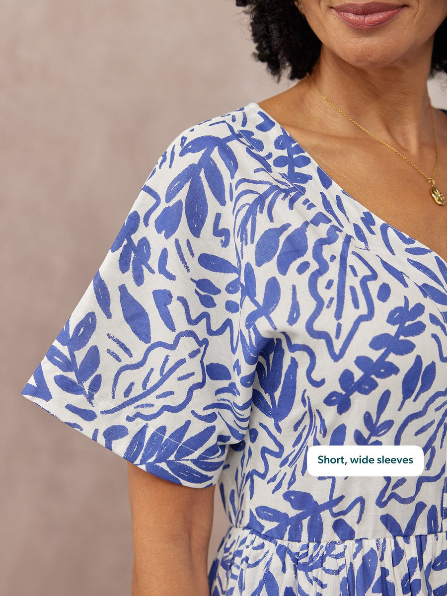 Close-up of a model wearing a white dress with a blue abstract print, short wide sleeves, V-neckline, and a tiered skirt.