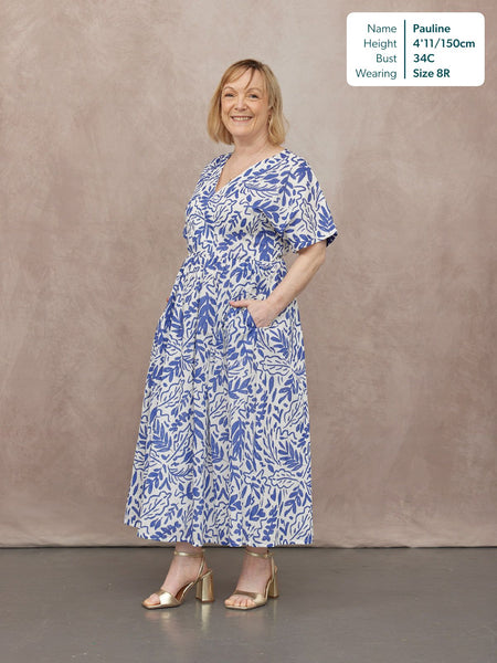 Model wearing a white midi dress with blue botanical print, short flutter sleeves, and a full skirt, styled with gold…