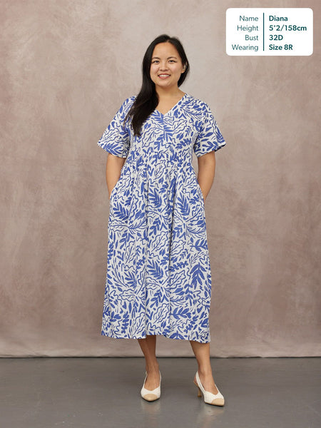 Model wearing a white midi dress with blue botanical print, flutter sleeves, V-neck, gathered waist, and side pockets…
