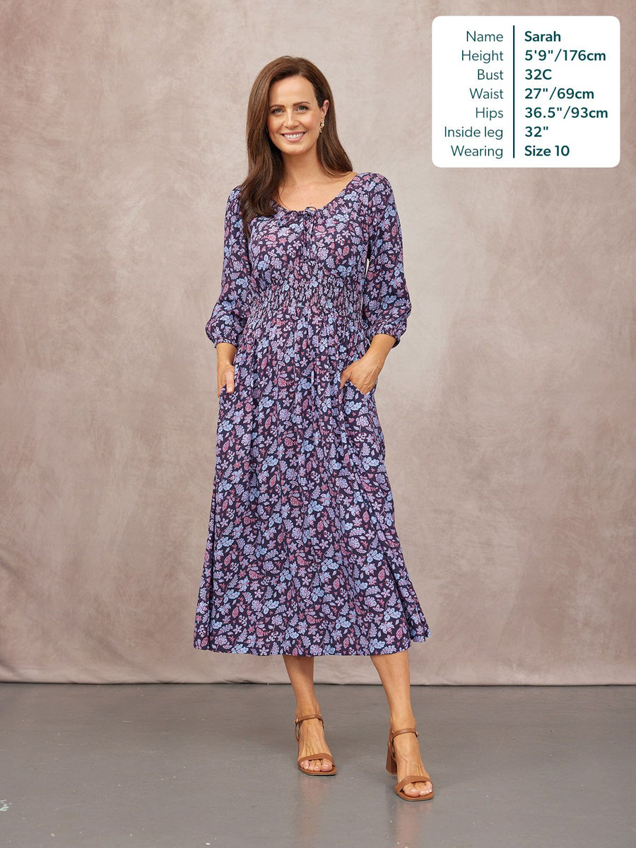 Model wearing a navy floral midi dress with a shirred waist, V-neck, 3/4 sleeves, and pockets, styled with tan block-heels