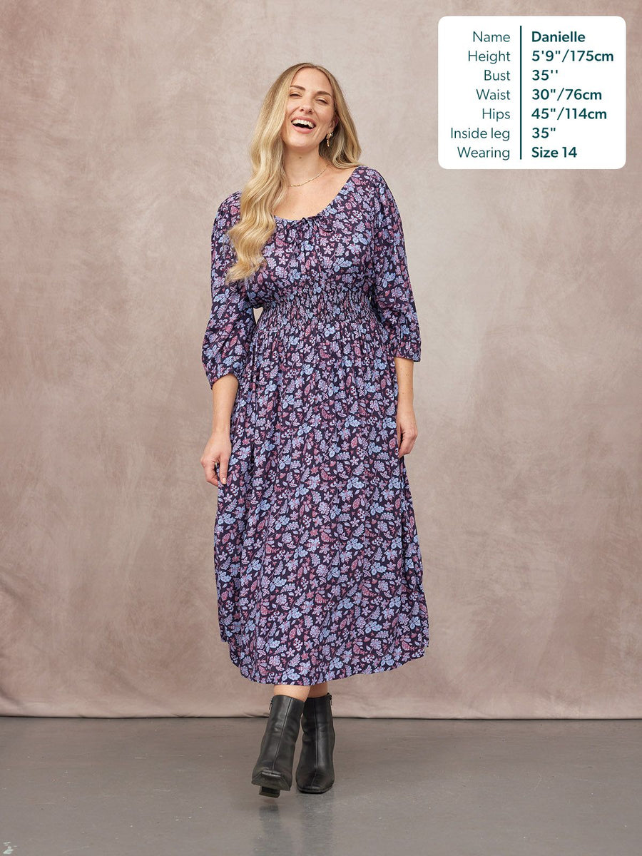 Model wearing a navy floral midi dress with shirred waist, scoop neck, and 3/4 sleeves, styled with black ankle boots.