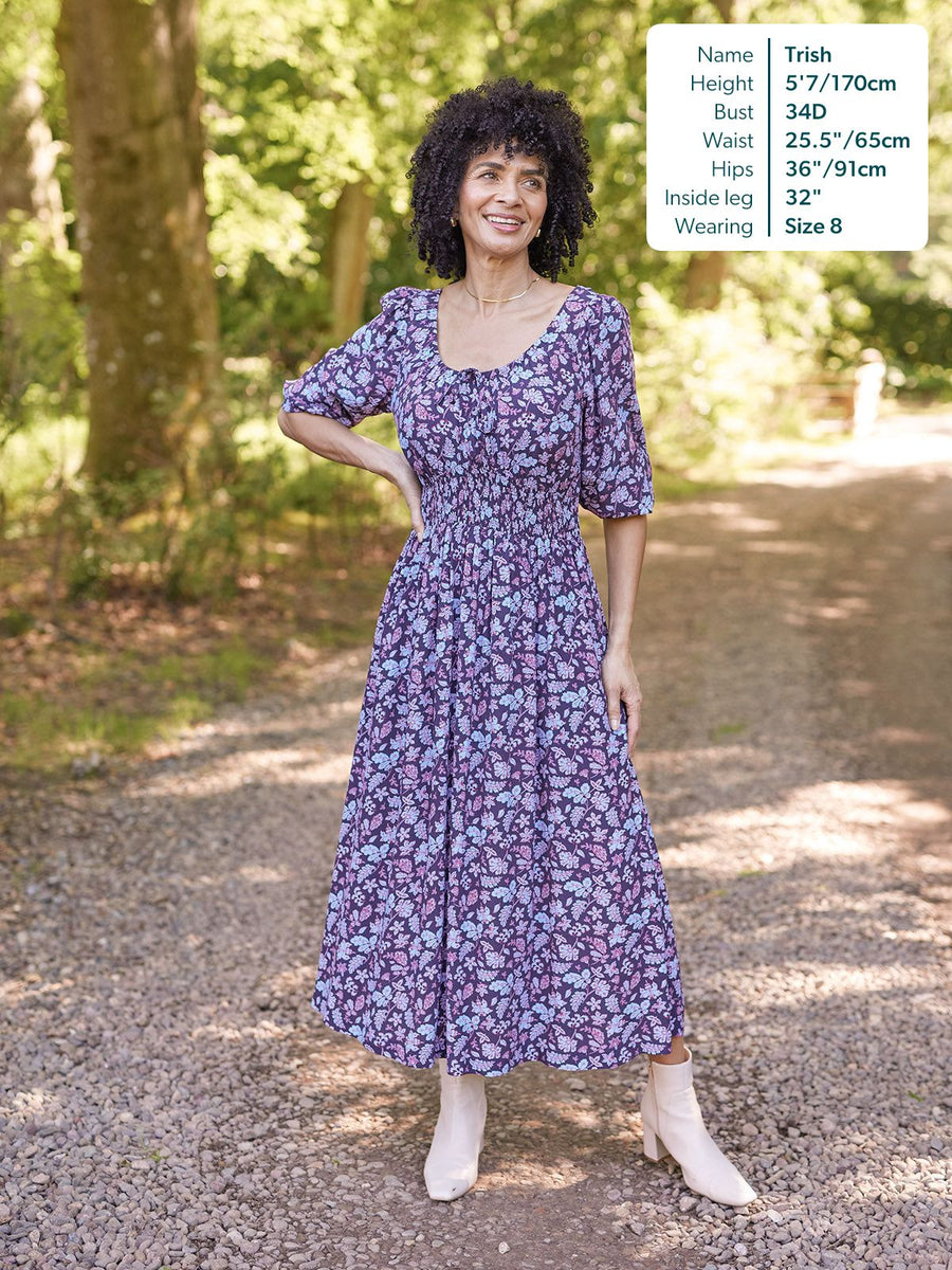 Model wears a purple floral midi dress with puff sleeves, shirred waist, and scoop neck, styled with white ankle boots