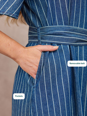 Close-up of a blue denim pinstripe dress with a tied waist belt and side pocket, model's hand resting in pocket.