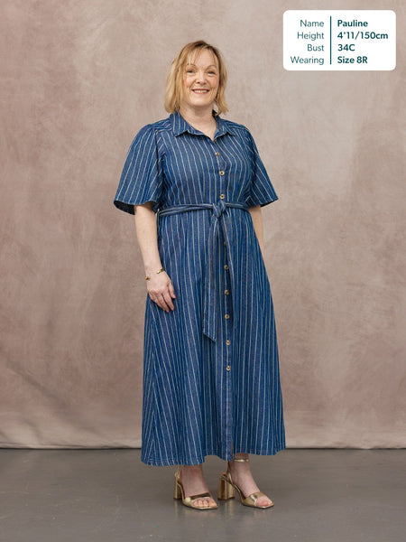 Model wearing a blue denim pinstripe midi shirt dress with short sleeves, button-through front, and a self-tie belt at the…