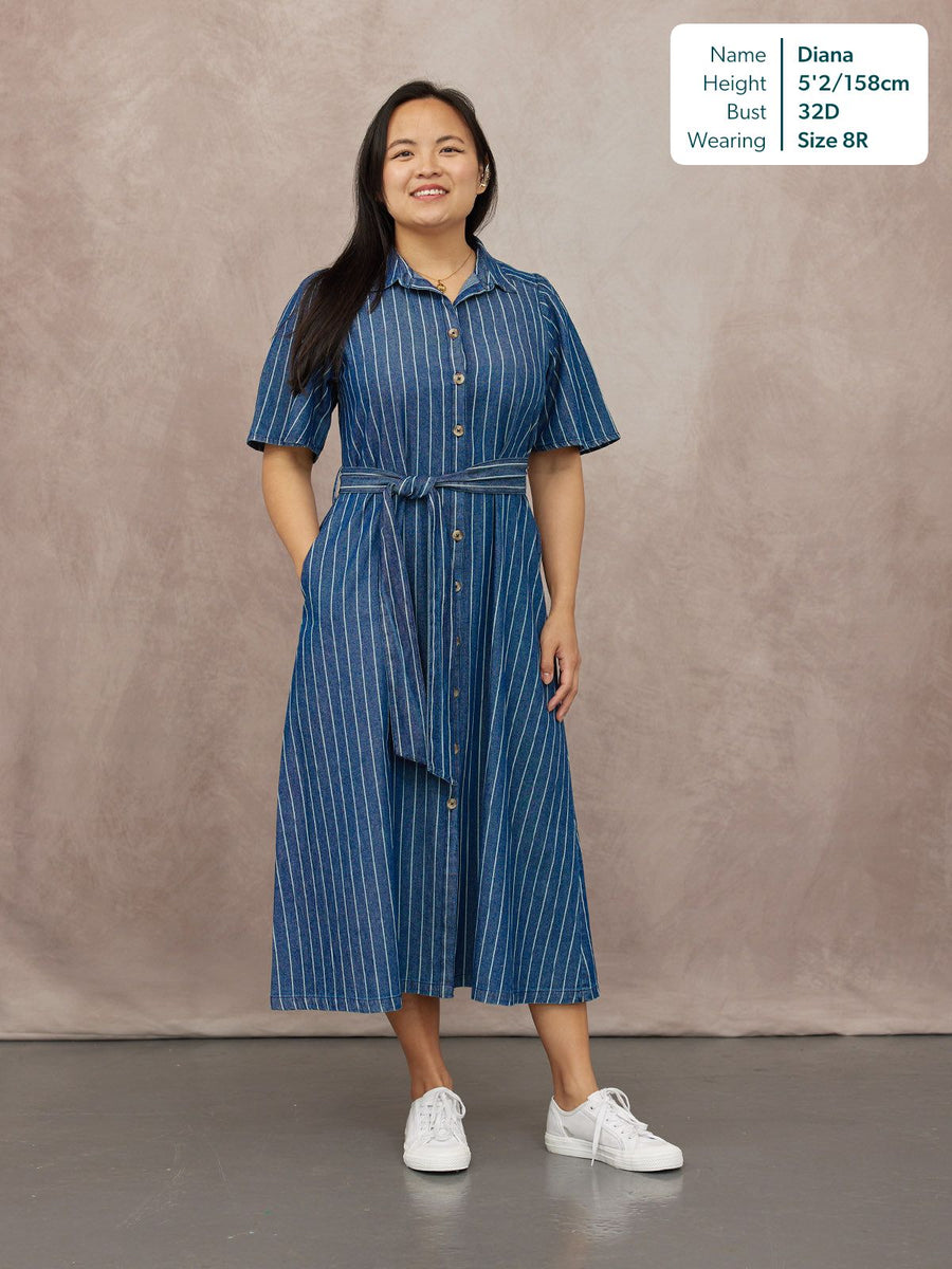 Model wears a blue denim pinstripe midi shirt dress with short sleeves, button front, and tie belt, styled with white…