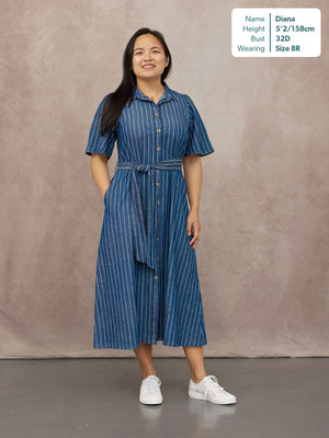 Model wears a blue denim pinstripe midi shirt dress with short sleeves, button front, and tie belt, styled with white…