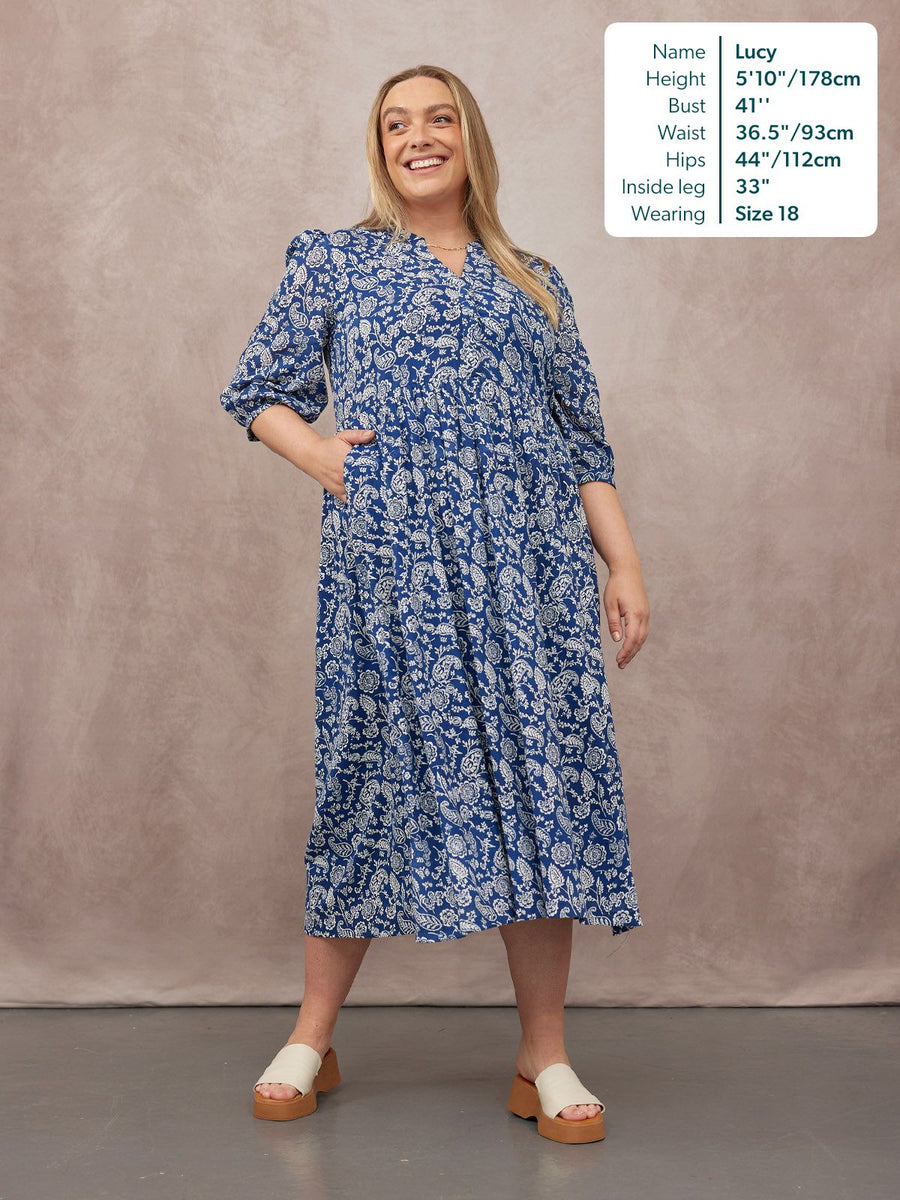 Model wears a blue and white paisley print tiered midi dress with puff sleeves and a V-neckline, styled with white platform…