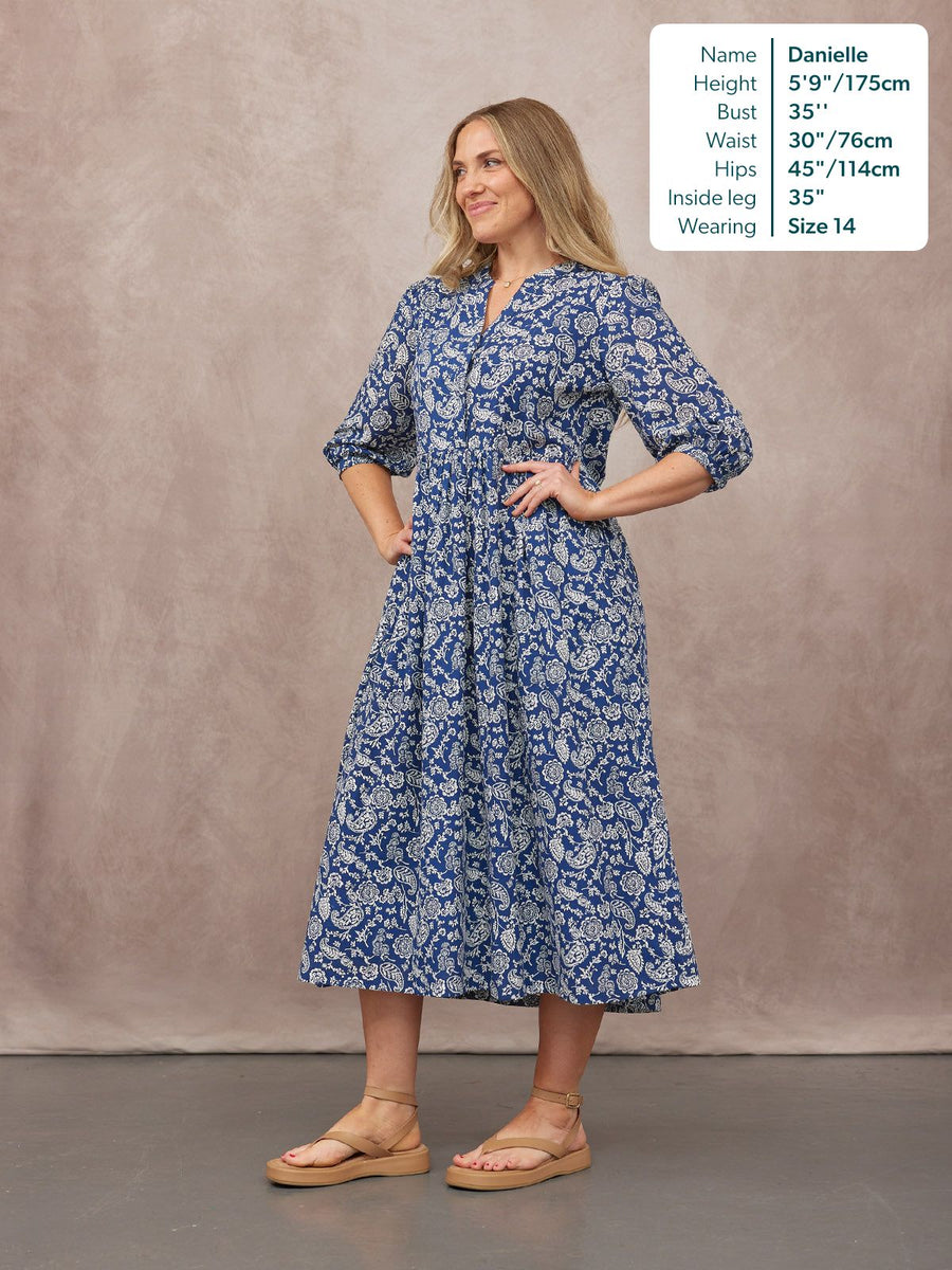 Model wears a blue paisley midi dress with 3/4 puff sleeves, V-neckline, and tiered skirt, paired with tan platform sandals.