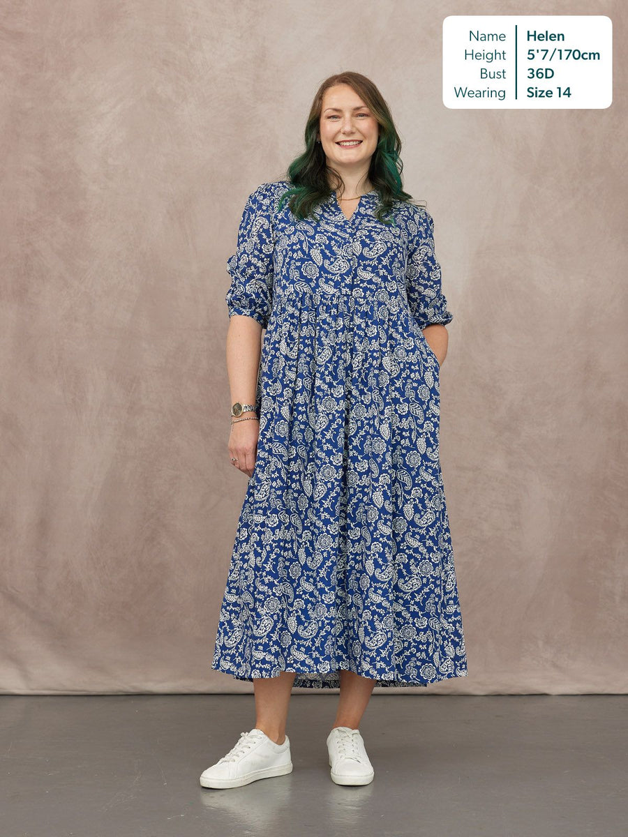 Model wears a blue and white paisley print tiered midi dress with 3/4 sleeves and a notch neckline, styled with white…