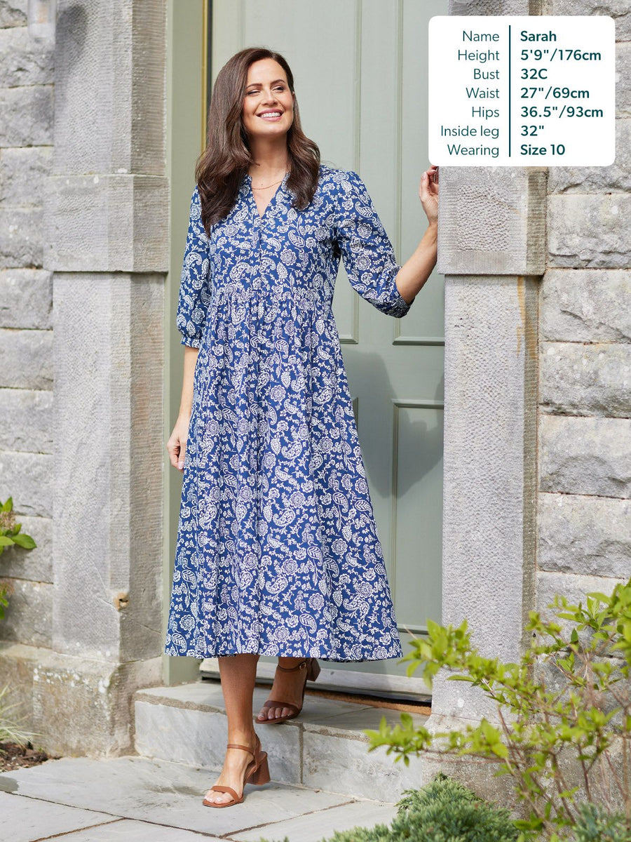 Model wears a blue and white paisley midi dress with V-neck, three-quarter sleeves, and tiered skirt, paired with tan…