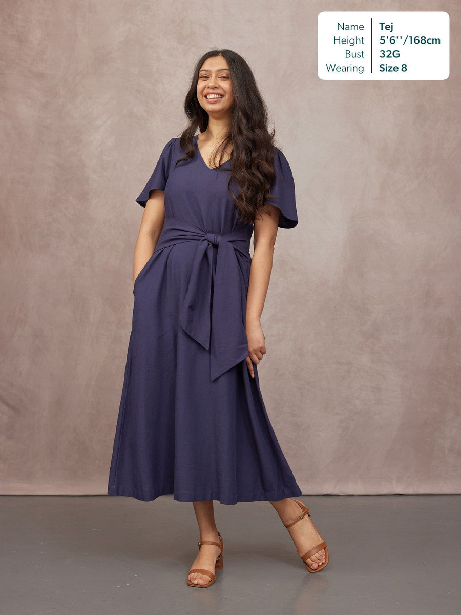 Model wearing a navy blue midi dress with flutter sleeves, V-neckline, tie waist belt, and tan block-heel sandals.