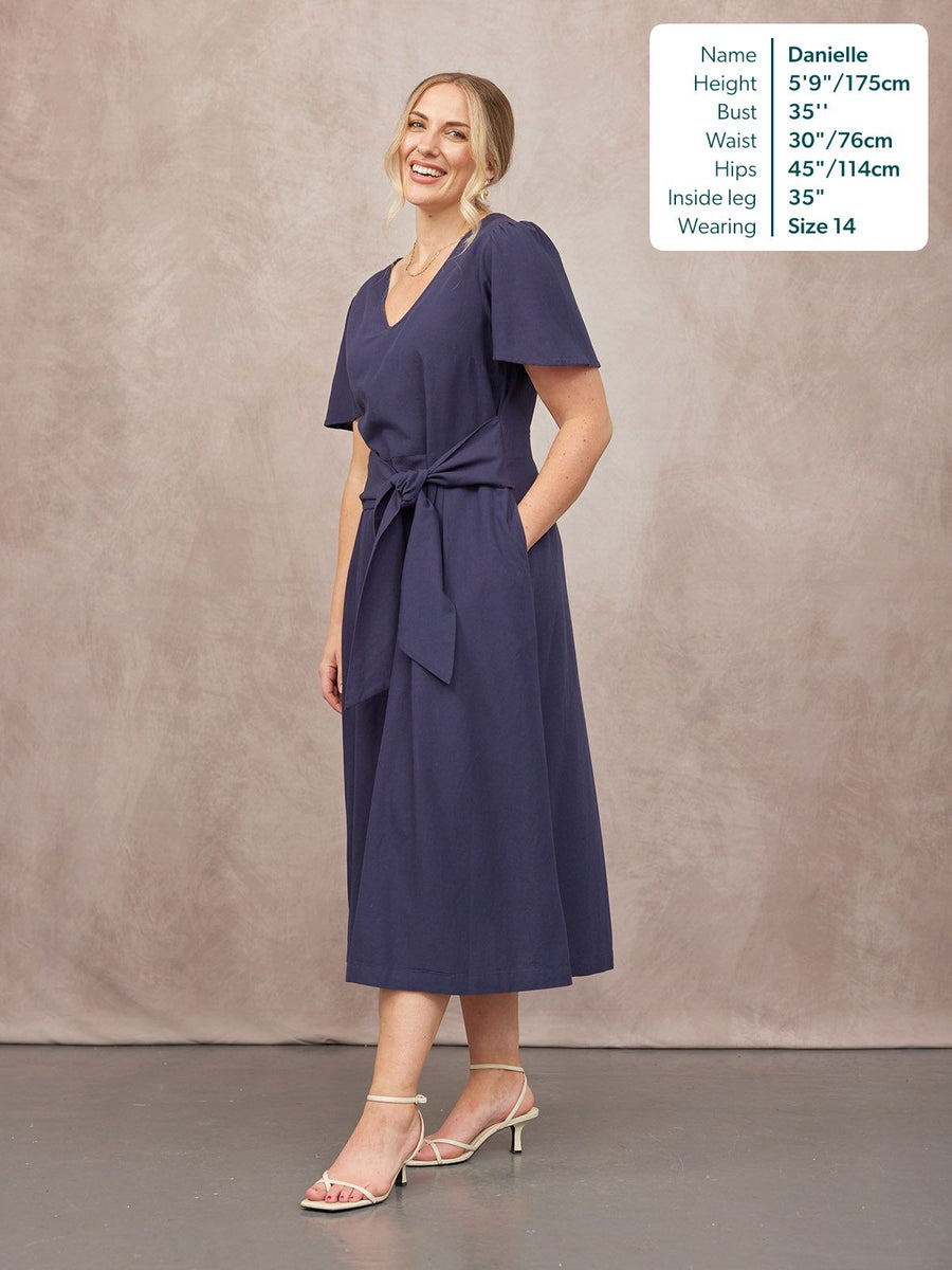 Model wears a navy midi dress with a V-neck, flutter sleeves, tie waist, and full skirt, styled with strappy cream heels.