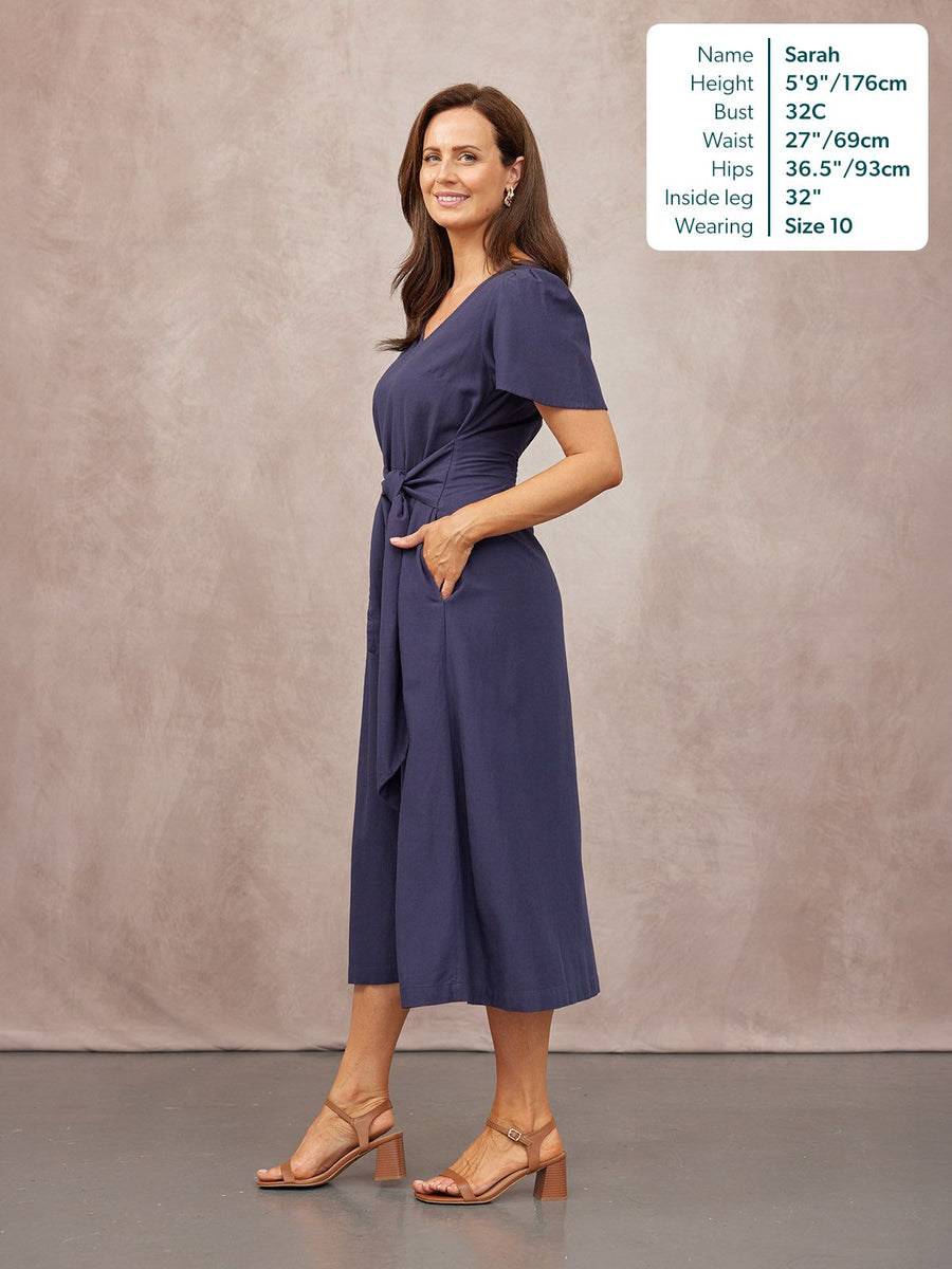 Model in a navy short-sleeve midi dress with a tie waist and side pocket, paired with tan block-heel sandals.