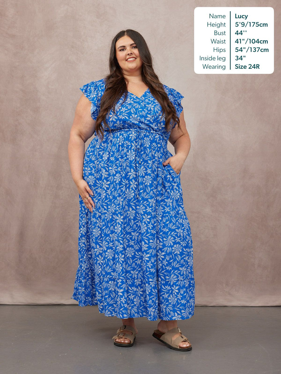 Model wearing a blue floral maxi dress with frill sleeves, V-neck, and tiered skirt, styled with tan sandals.