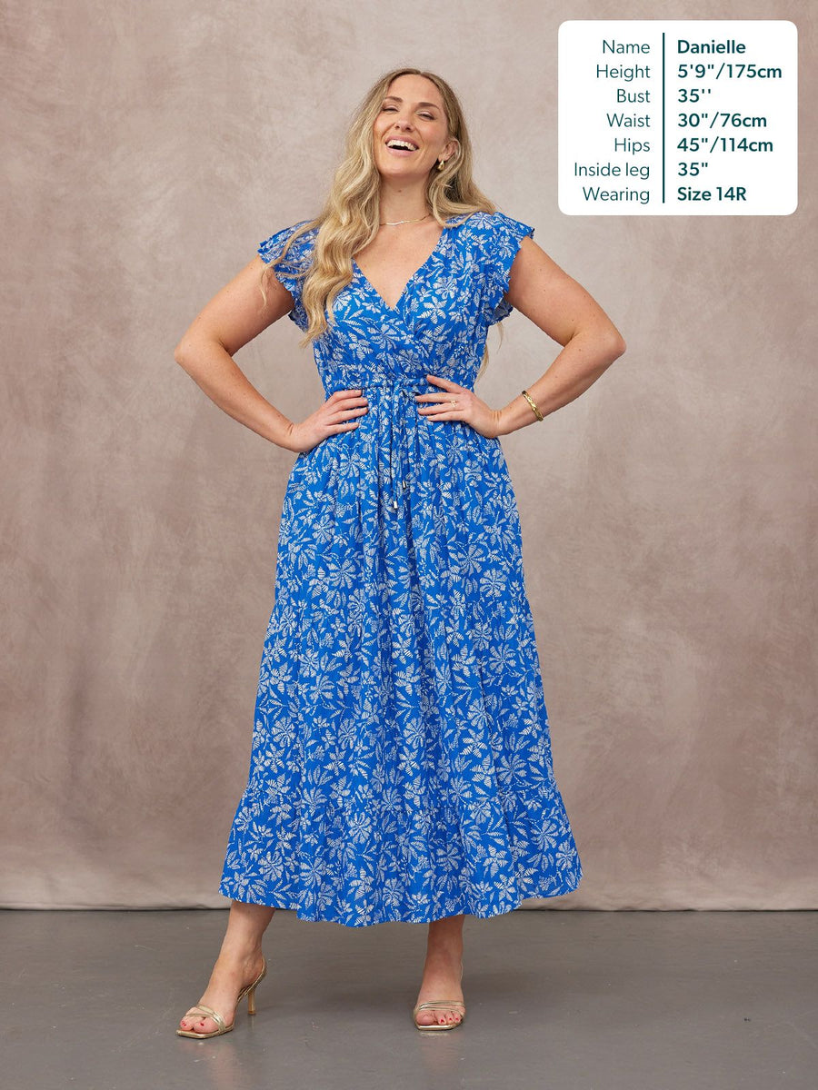 Model wearing a blue and white floral maxi dress with flutter sleeves, V-neckline, and tie waist, paired with gold heels.