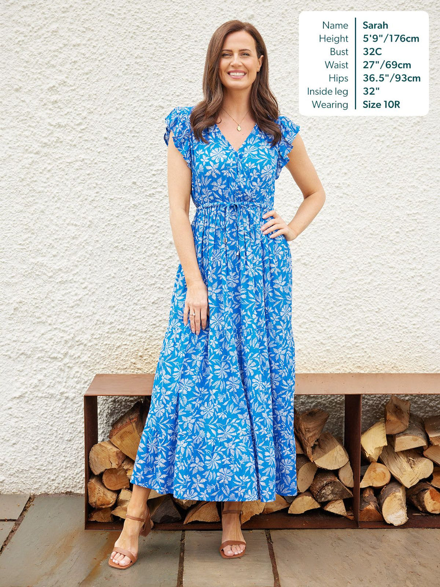 Model wearing a blue floral maxi dress with ruffle sleeves, V-neck, and drawstring waist, styled with tan heeled sandals.