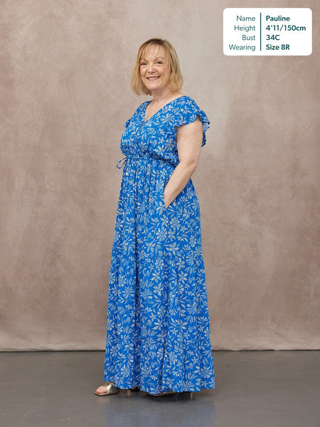 Model wearing a blue floral maxi dress with flutter sleeves, V-neck, drawstring waist, and tiered skirt, styled with gold…