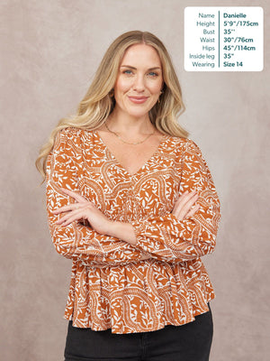 Model wears a rust orange and white floral print V-neck top with long sleeves and a peplum hem, paired with black trousers.