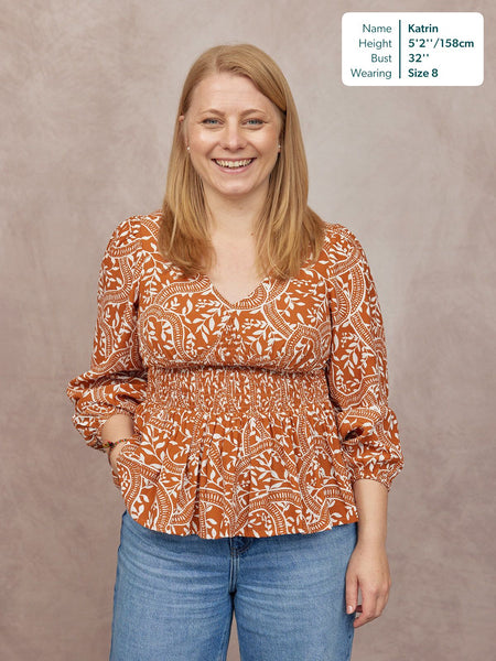 Model wearing a rust orange and white paisley print V-neck top with smocked waist and blouson sleeves, paired with blue…