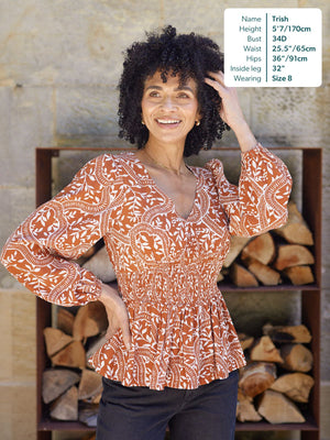Model wearing a rust orange floral print top with a V-neck, puff sleeves, and smocked waist, paired with dark jeans outdoors.