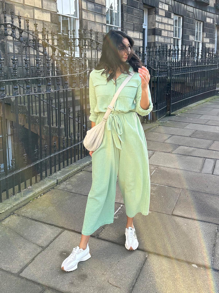 A woman wearing the Clara jumpsuit paired with trainers and a cross-body bag, pictured walking along the street in the sunshine. 