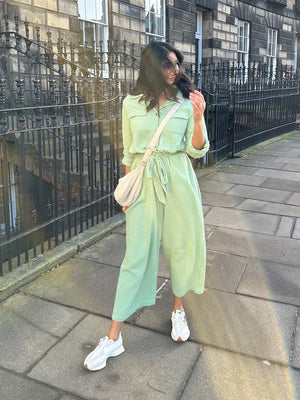 A woman wearing the Clara jumpsuit paired with trainers and a cross-body bag, pictured walking along the street in the sunshine. 