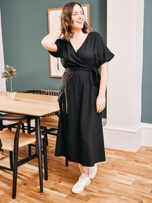 A model stands in a dining area wearing the Daisy wrap dress and white trainers, pictured looking off to the side and laughing. 